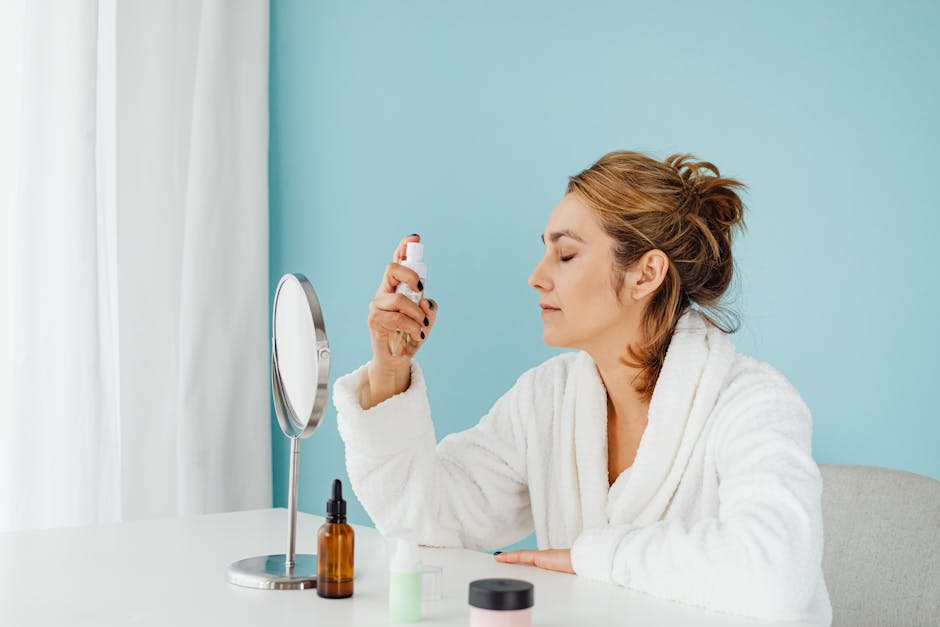 Woman in a bathrobe using a skincare spray in front of a mirror indoors