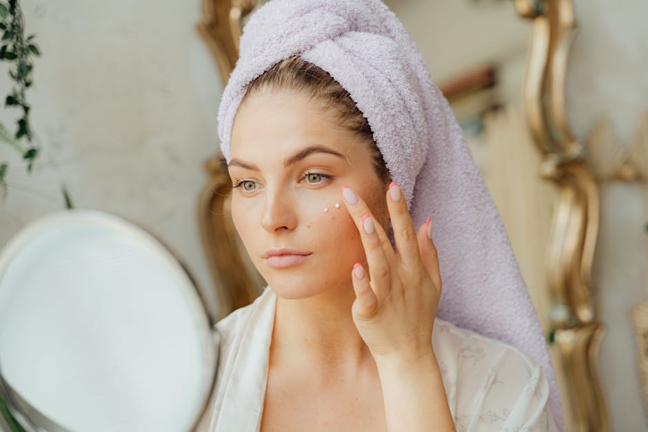 Woman with head towel applying moisturizer in front of mirror indoors
