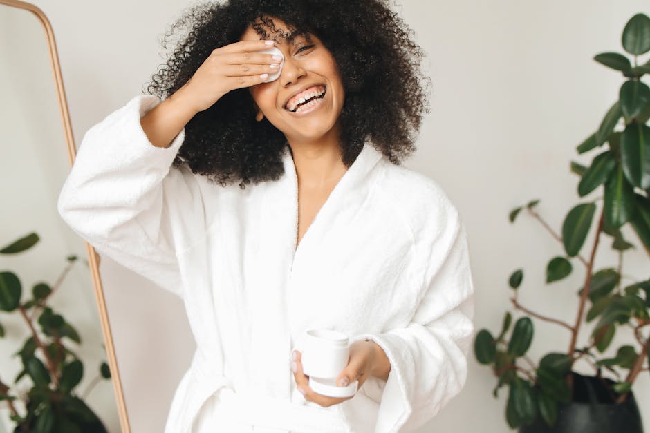 Happy woman in bathrobe applying skincare routine with cotton pad indoors