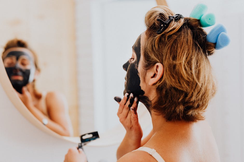 A woman is applying a black facial mask in front of a mirror, focusing on skincare and beauty routines